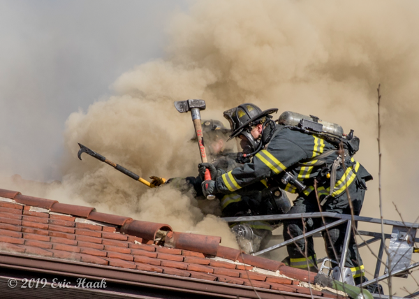 dramatic photo of Firefighters venting a roof in heavy smoke from aerial ladder