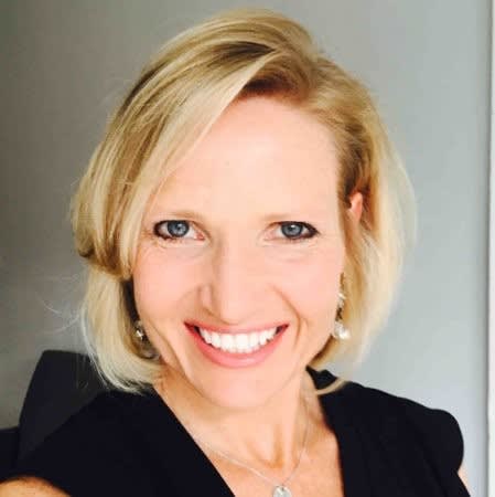 Headshot of blog author Julie Kae. She has short blonde hair and blue eyes and smiles at the camera, wearing a black top and a necklace, with a plain light-colored background.