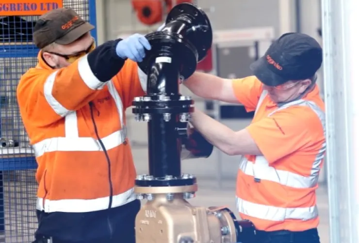 Two workers in safety gear and high-visibility clothing install or repair industrial piping in a facility. One holds a pipe vertically while the other secures it.