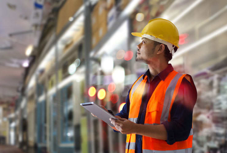 A construction worker in a yellow hard hat and orange safety vest holds a tablet while inspecting an indoor building site.