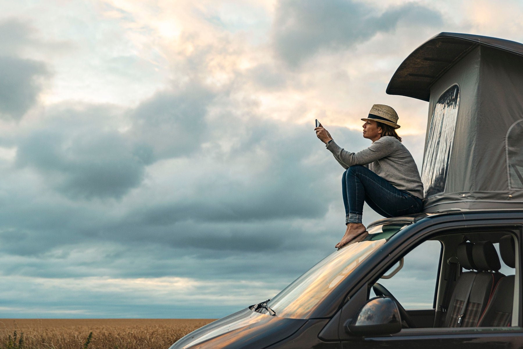 Woman taking a photo whilst sitting on roof of camper at sunset