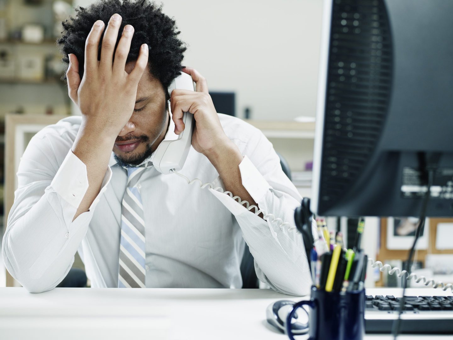 A man in a white shirt and tie sits with elbows on his desk; one hand is holding the phone to his ear and one hand covers his face.