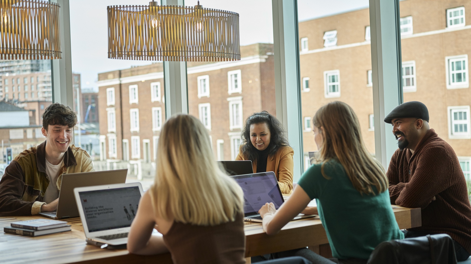 A group of five, collaborating around a desk with their laptops chat together.