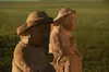 The image shows two ancient stone statues set against a backdrop of a grassy field, possibly at sunset or sunrise. The statues appear to be weathered and eroded, suggesting significant age. Each statue depicts a figure wearing a hat, and both are seated with hands resting on their knees or in a meditative pose.