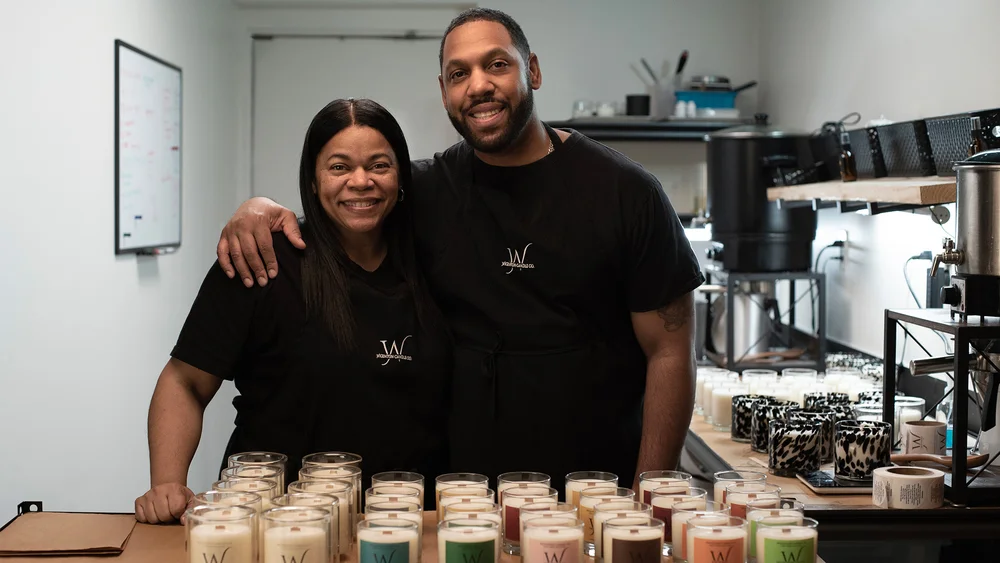 A man and a woman stand in a workshop in front of an array of candles