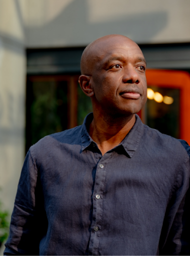 A bald Black man wearing a long sleeve navy buttoned up shirt stands, posted against a wall