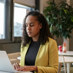 A woman in a yellow blazer works on her laptop