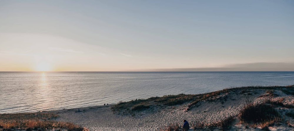 Fotografía de una puesta de sol sobre el océano con dunas de arena y personas en la playa