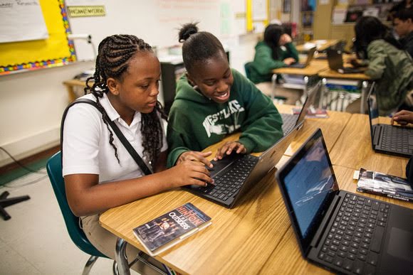 Zwei Schülerinnen einer weiterführenden Schule sitzen in einem Klassenzimmer und teilen sich einen Laptop. Das Mädchen im weißen Polohemd sieht konzentriert auf den Bildschirm. Ihre Freundin im grünen Kapuzenpullover schaut lächelnd auf den Laptop.