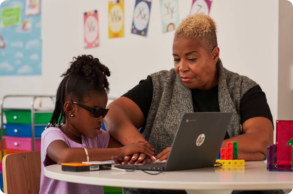 Una alumna joven con discapacidad visual usa una Chromebook con la ayuda de un lector braille mientras la profesora, sentada a su lado, le guía la mano.