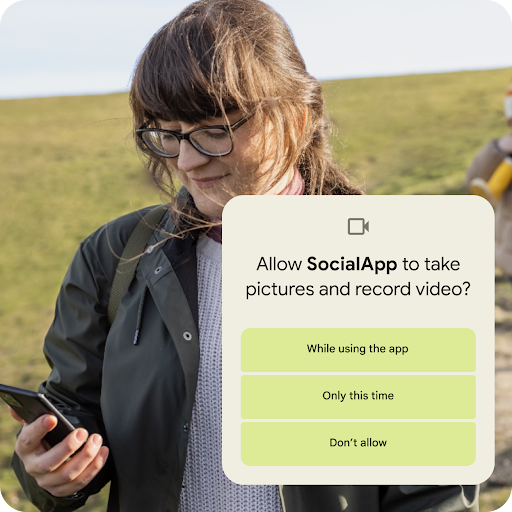 A person standing on a grassy hillslope looking at their Android phone. A graphic overlay seeking permission to allow an app to take pictures and record videos is placed on top. Permission options include allowing access while using the app, only this time, or do not allow.