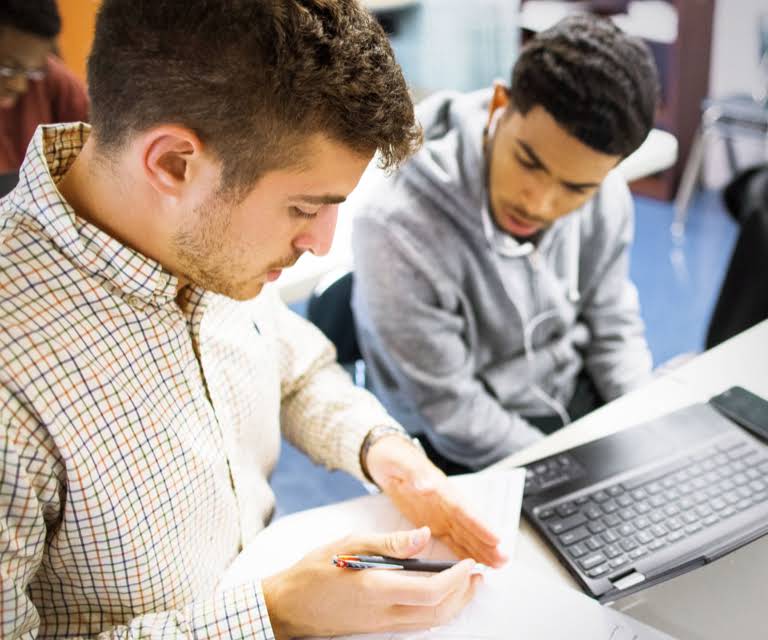 One student looks like they are explaining something on a sheet of a paper to another student.