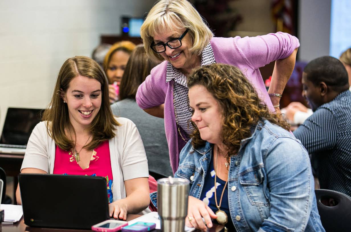 Três docentes sorriem enquanto olham para um laptop. Eles estão em uma sala de conferências cheia de pessoas sentadas ao redor de mesas e trabalhando em laptops.