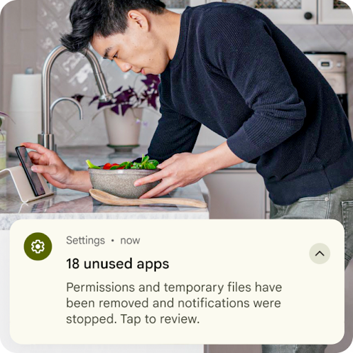 Một người vừa chuẩn bị đồ ăn bên bồn rửa vừa xem điện thoại Android. Lớp phủ đồ hoạ phía trên hình ảnh này là thông báo liên quan đến chế độ cài đặt. Thông báo cho biết các tệp tạm thời từ những ứng dụng không dùng đến đã bị xoá và các quyền đã được đặt lại.