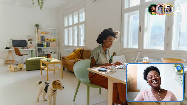 Duas mulheres conversam por videochamada em uma reunião de equipe. Uma delas está à mesa e tem um cachorro por perto. A outra está sorrindo na janela da videochamada.