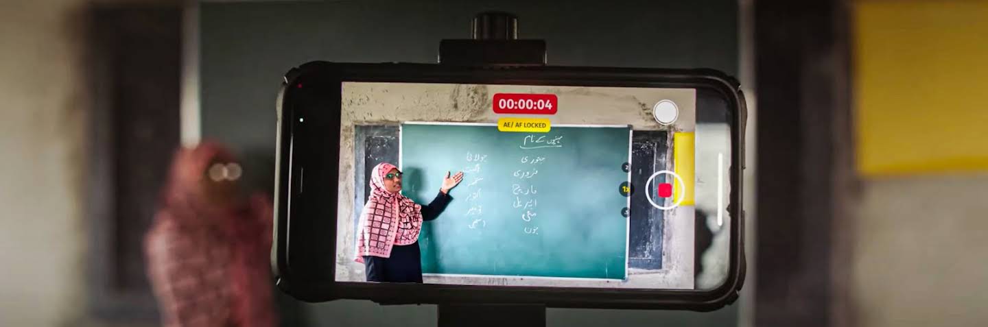 A person sitting next to a crate of eggs. In their left hand is a phone displaying a teacher in front of a chalkboard. In their right hand is a pen resting on a notebook.