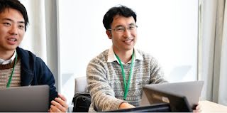 Two men, one wearing glasses, are having a conversation and smiling while working on laptops.