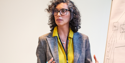 A woman in a yellow blouse and blazer speaks to a crowd