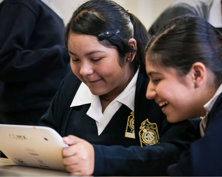 Duas adolescentes de uniforme estão sorrindo, e uma delas está segurando um tablet