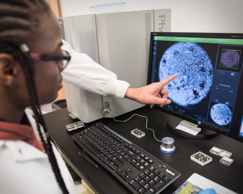 Finger pointing to a computer screen with a woman looking closely. It looks as if they are in a science lab and they are looking at a close up of a circular mass.