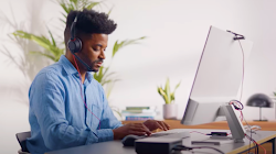 Een persoon die een microfoonheadset en een blauw overhemd draagt, zit aan een bureau en typt op een desktop.