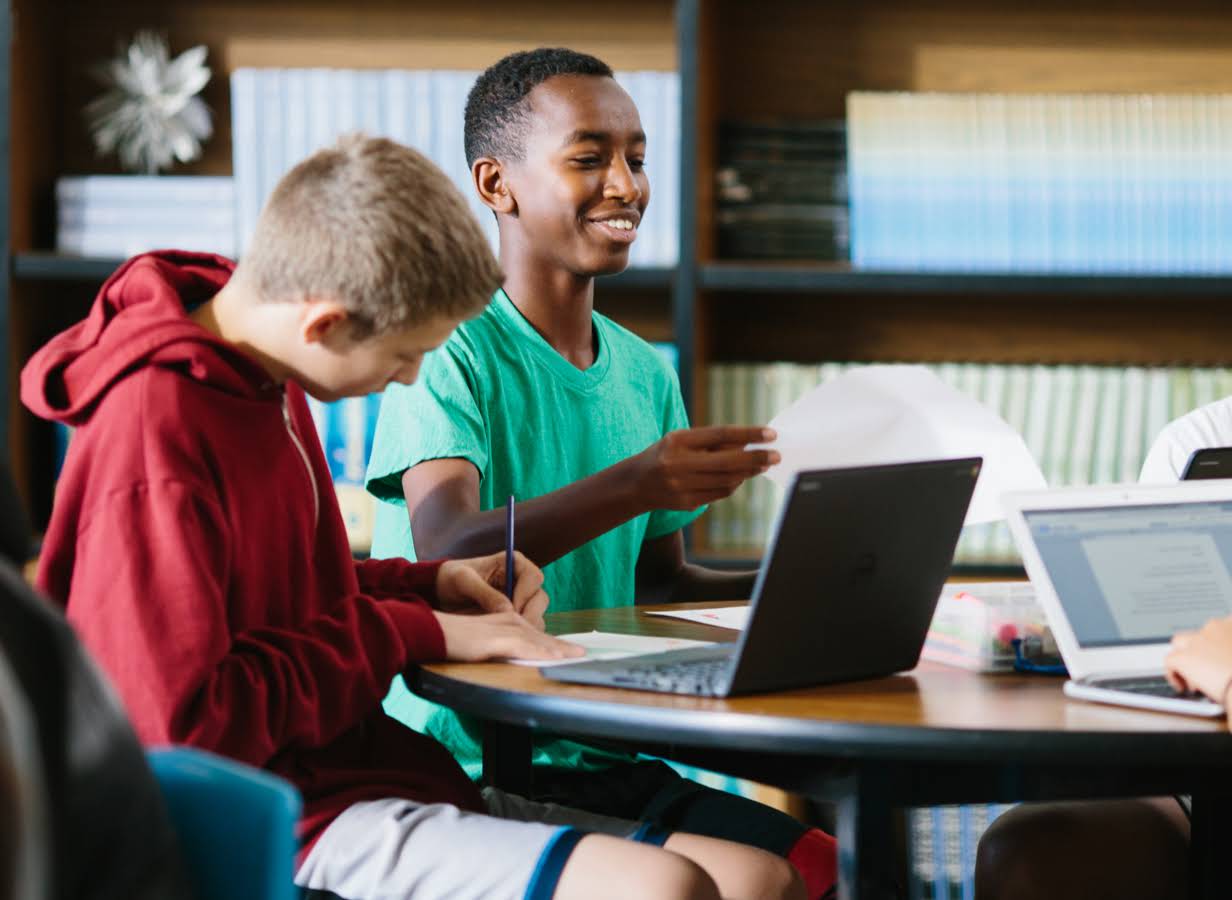 Seorang siswa memegang selembar kertas dengan siswa lain yang mengerjakan tugas di dekatnya menggunakan Chromebook.