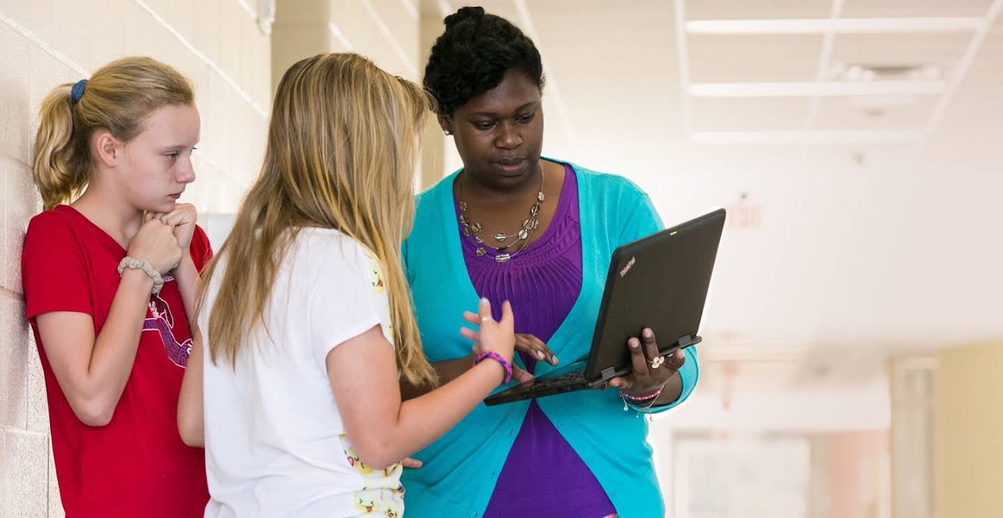 Adult educator shows something on a laptop screen to two students in a hallway.