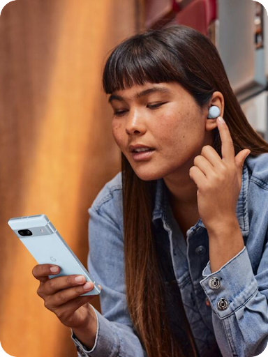 An Android user is holding a conversation, touching their earbud with their finger, and looking down at their Android phone.