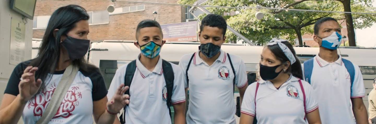 En una sala de reuniones, dos personas con mascarilla que están sentadas a una mesa frente a frente miran las laptops que tienen abiertas. Sobre la mesa, hay dos laptops más y un cuaderno.