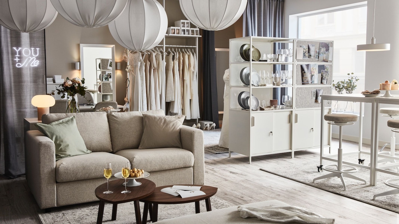 A wedding planner showroom with a sofa and coffee table, a shelving unit displaying tableware, and a rail with white dresses.