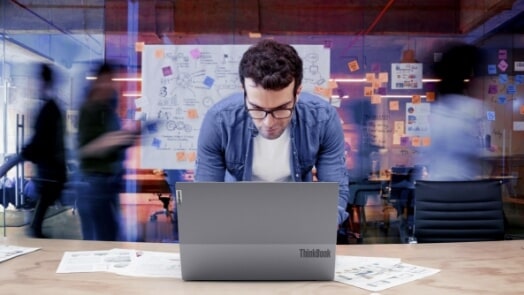 Man working on a laptop as people walk behind him in a busy, creative office