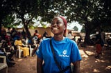 Photo of Ruth Alanana, a 25-year old “Data Advocate” for the Data Made Simple project. Ruth is standing outside wearing a blue t-shirt with text “Data Made Simple.” Ruth is standing outside in a rural setting of Nigeria, smiling, and looking off camera. In the background are the backs of people seated in rows.