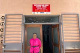 Esnart is wearing a bright pink blouse, and is standing outside of the maternal and child health ward, which is a beige pink building with a bright red sign.