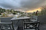 A grey table on a balcony showing a pretty sunset vista of hillside homes