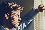 Pensive young man gazes out window. The blinds cast striped shadows on his face.