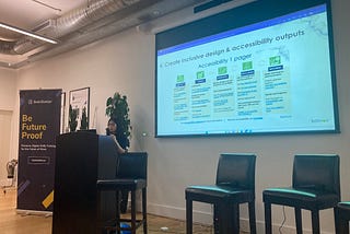 Christina Lai, an east asian woman of short stature presents inclusive design and accessibility outputs on a projector, next to podium and 3 empty stools. Banner beside her reads ‘BrainStation. Be Future Proof’