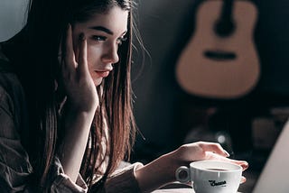 A young woman with long brown hair staring at her laptop while drinking coffee. She looks slightly perplexed.