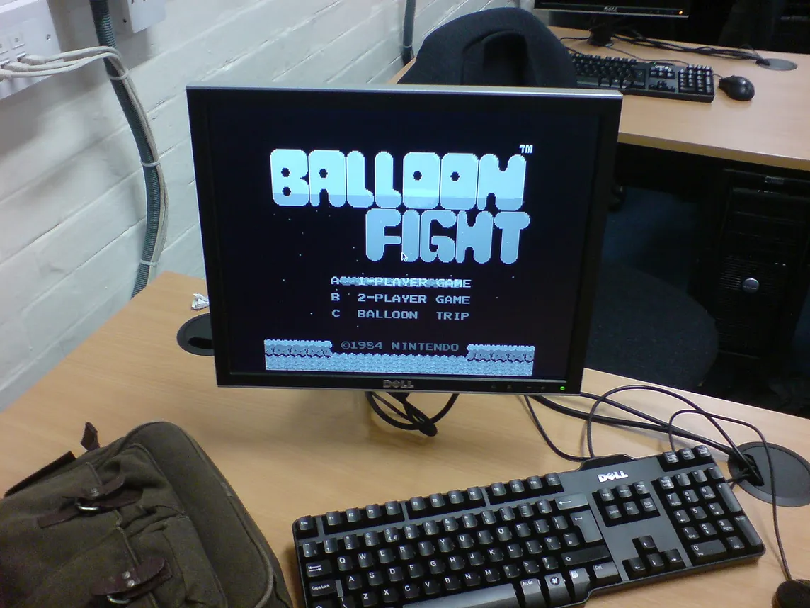 A computer screen on a desk with keyboard and mouse showing the title screen of the game “Balloon Fight” in black and white.