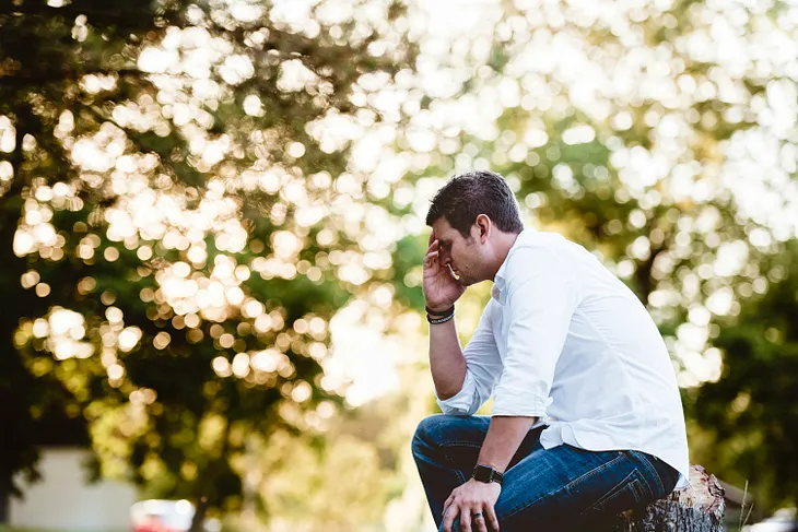 A man sitting outside with a hand to his head thinking deeply. The branches of the trees play with the background light in unsharp figures