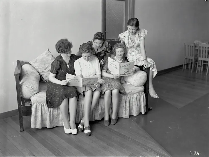 Black and white photo of five women sitting on a couch together reading the newspaper for job oppportunities.