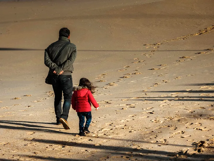 A color photo of a father and young girl with backs to viewer weariong parkas walking on a beach.