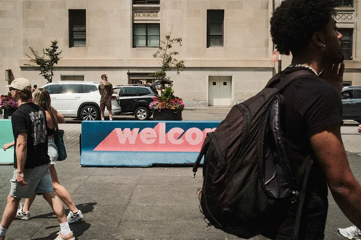 Pedestrians moving along the street, with a colored concrete barrier in the center of the frame that reads, “welcome.”