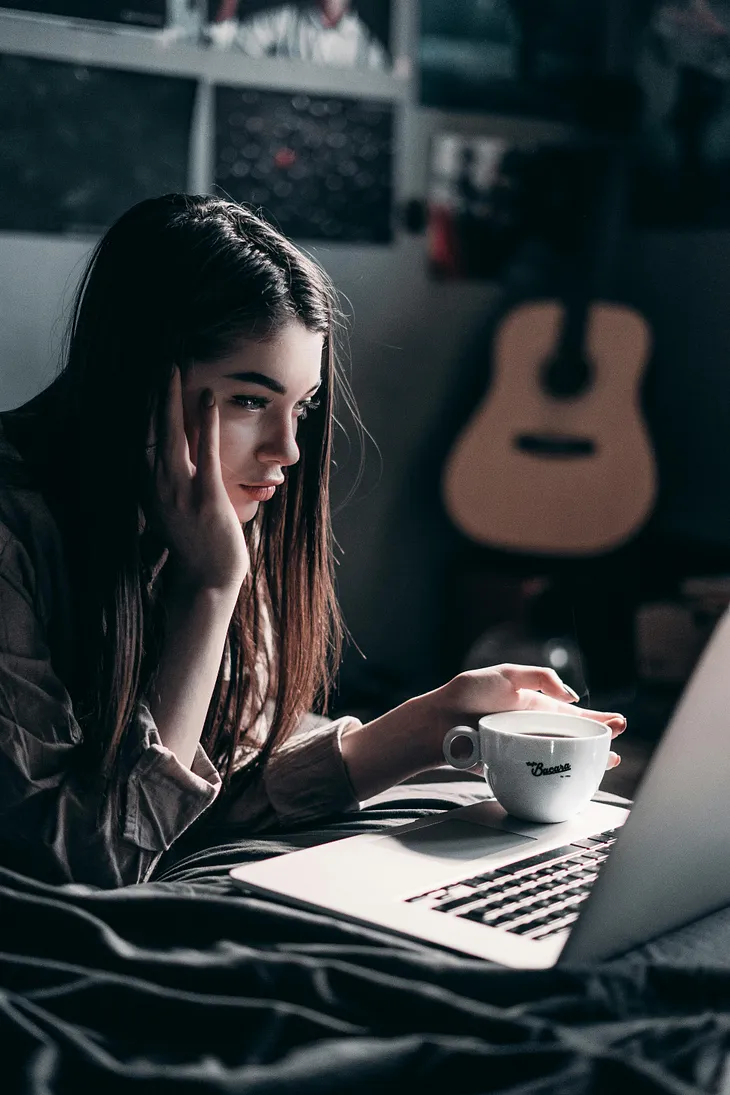 A young woman with long brown hair staring at her laptop while drinking coffee. She looks slightly perplexed.