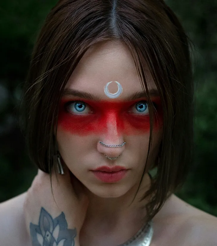 A woman with blue eyes, a nose piercing, and red eye makeup poses in a front-on image. She has a white decorative symbol on her forehead.