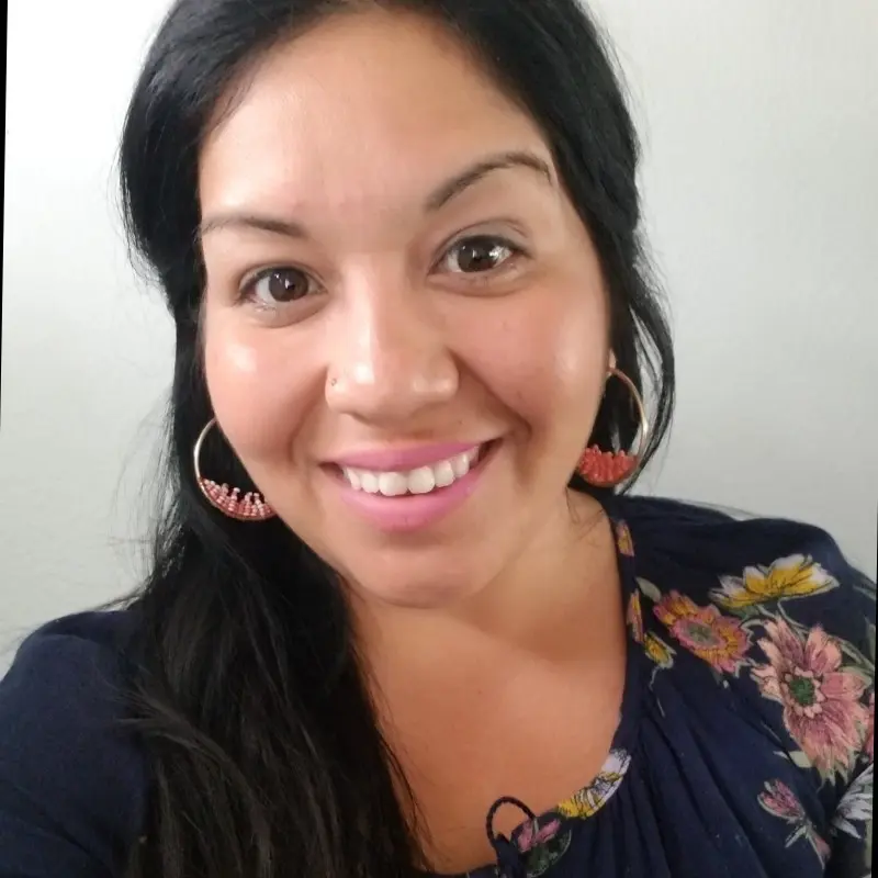 A headshot of Monica Jayasighe, who is wearing a black floral shirt and smiling in front of an off-white background.