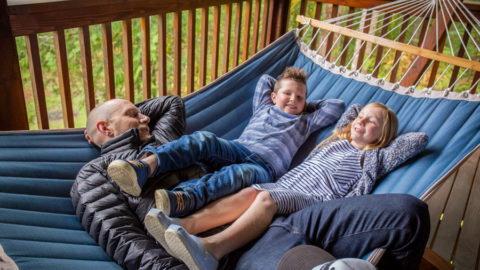 A man lays on a hammock with his children, a boy and a girl, who hang their legs over his lap