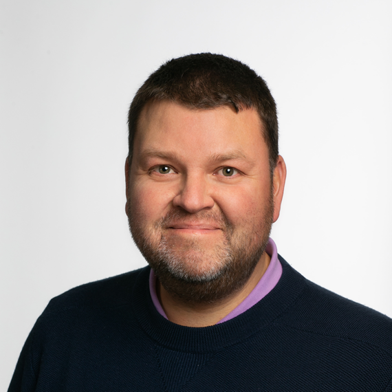 A man with short dark hair and a beard wearing a black sweater over a light purple collared shirt smiles at the camera against a plain white background.