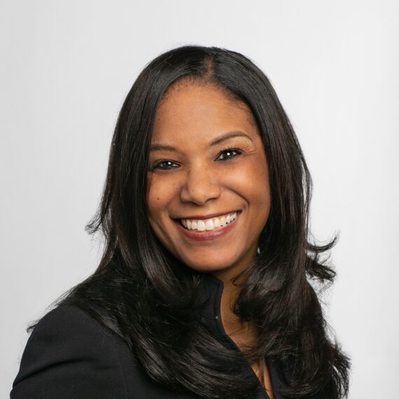 A woman with long black hair wearing a dark blazer smiles warmly in front of a plain white background.