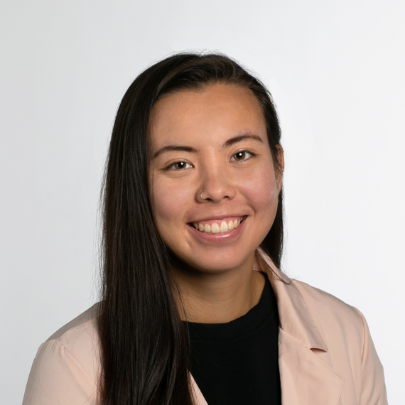 A person with long straight hair, smiling, wearing a light-colored blazer over a black top, against a plain white background.