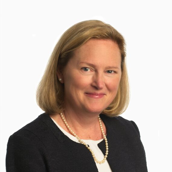 A woman with shoulder-length blonde hair wears a black blazer, white blouse, and pearl necklace. She is smiling slightly and facing forward against a plain white background.